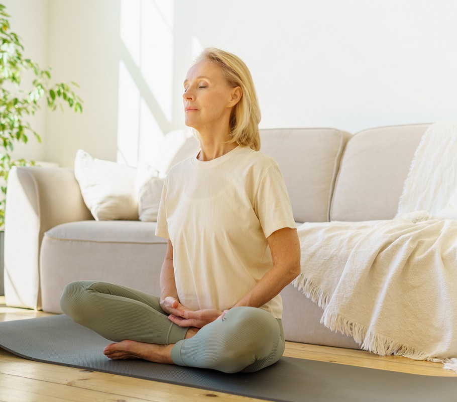 Peaceful senior woman in lotus position meditation with closed eyes at home while sitting on yoga mat on floor, full length.